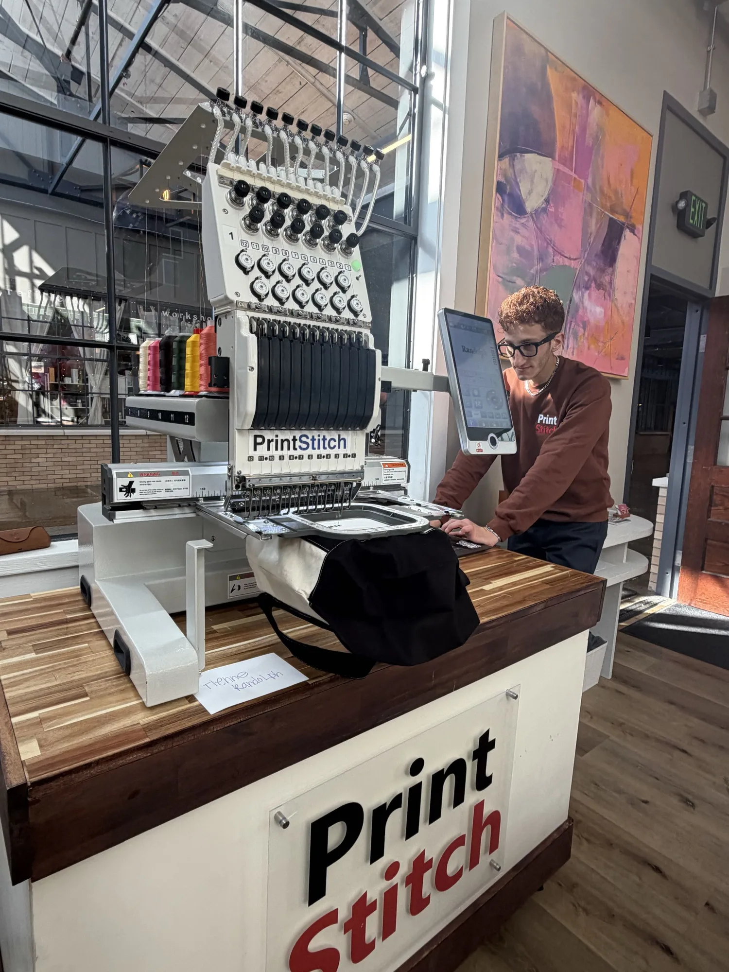 A PrintStitch embroidery machine on the branded counter at the MVMT House event, with a tote hooped under the needles