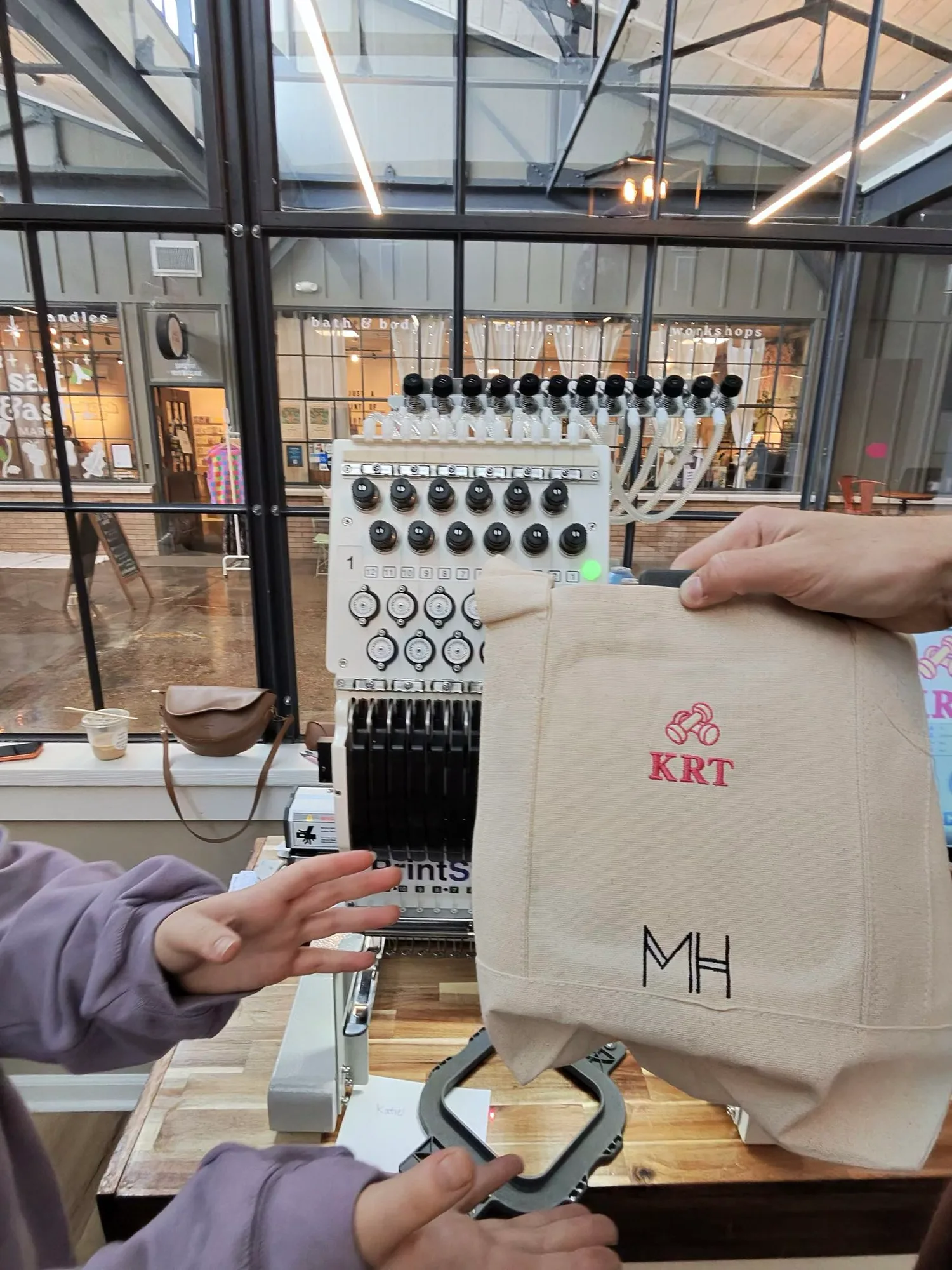 A canvas tote embroidered with a red dumbbell icon above the initials KRT, and MH stitched in black below